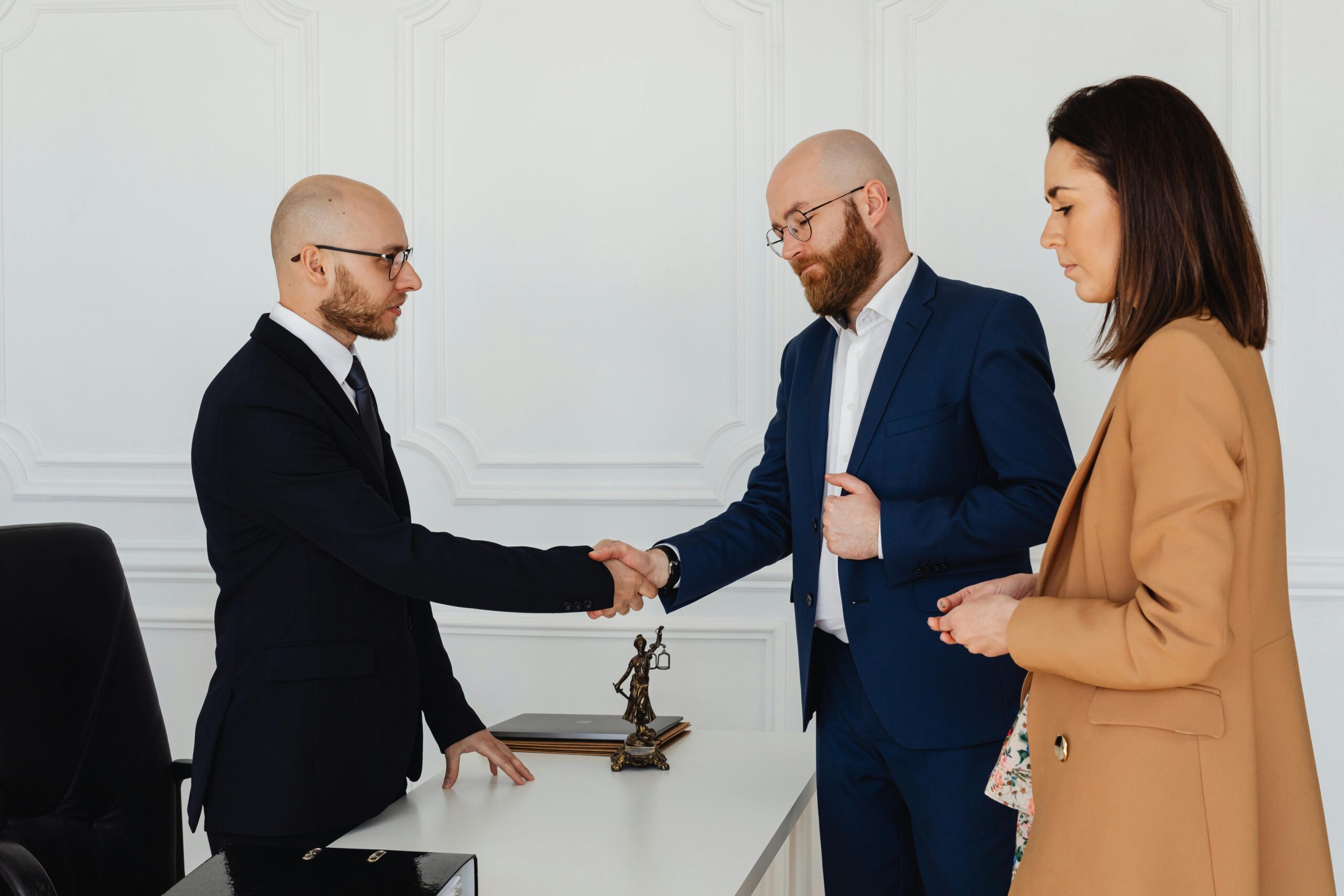 Three business professionals in formal attire shake hands during an office meeting.
